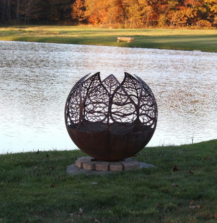 A metal sphere sculpture sits on the grassy edge of a pond.